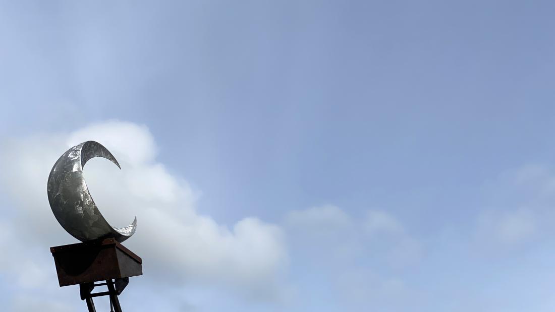 A crescent moon sculpture raised by a tractor arm against a blue sky.