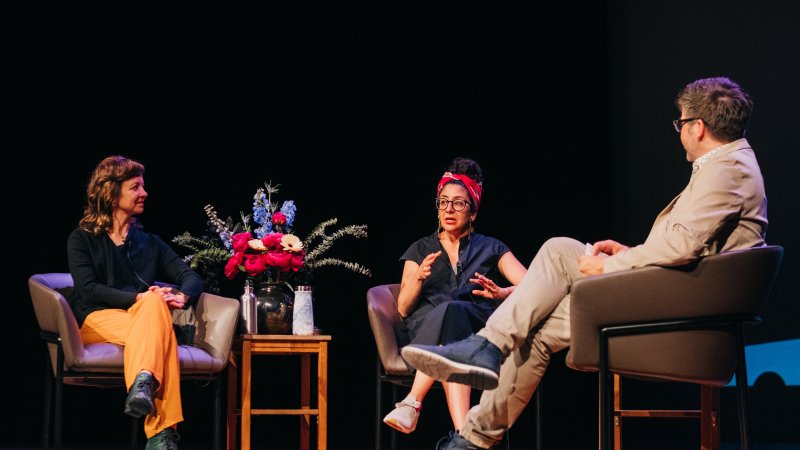 Artists Jaimie Robson and Maya Ersan sit on chairs on a stage, talking about their exhibition. Curator Jordan Strom sits across from them as moderator. A vase of flowers is placed on a table between the two artists.