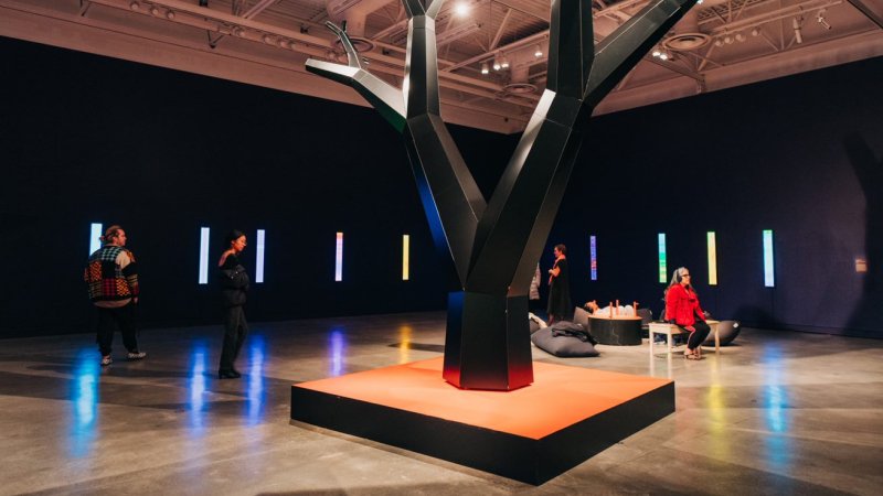Wood and metal sculpture of a forked tree on an orange square base by Charles Campbell in a purple gallery space, with gallery visitors nearby