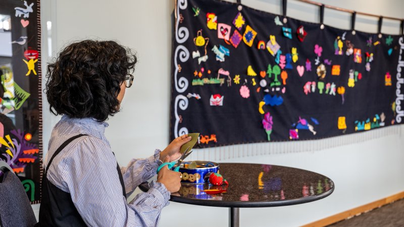 A woman sits next to a felt blanket hanging on the wall.