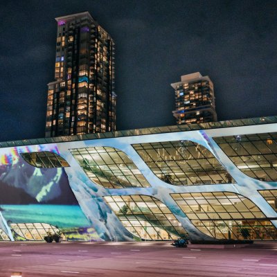 A projected artwork of blues and whites is shown on the exterior of Surrey Central Library at nighttime, which is lit up inside. A tall tower stands behind it.