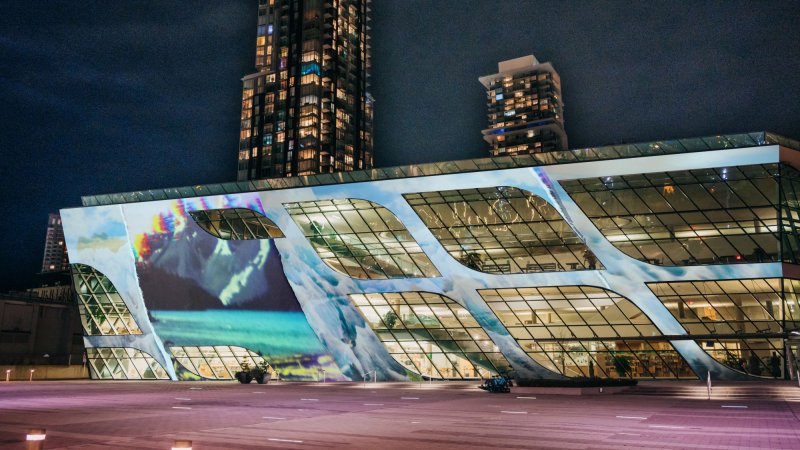 A projected artwork of a mountain landscape is shown on the exterior of Surrey Central Library at nighttime, which is lit up inside. A tall tower stands behind it.