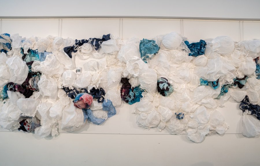 A textured wall art installation made of a variety of white and colored fabric scraps, arranged to create a dynamic, wave-like pattern.
