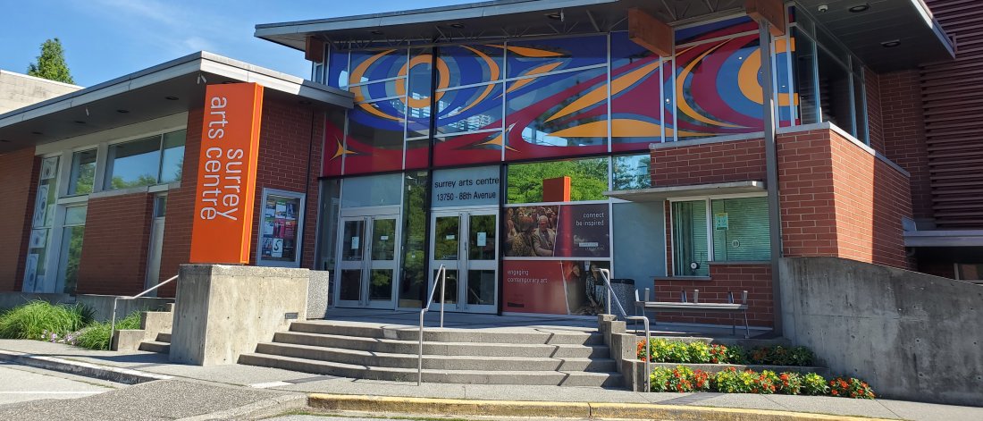 Surrey Arts Centre on a sunny day with blue sky. A colourful mural by Atheana Picha is on the top windows of the entrance. An orange rectangular sign says "surrey arts centre" in white letters next to the stairs leading to the doors.