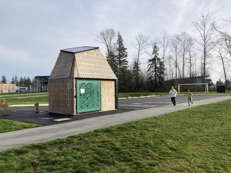 A small wooden structure with artistic designs, situated in a park with people jogging nearby.