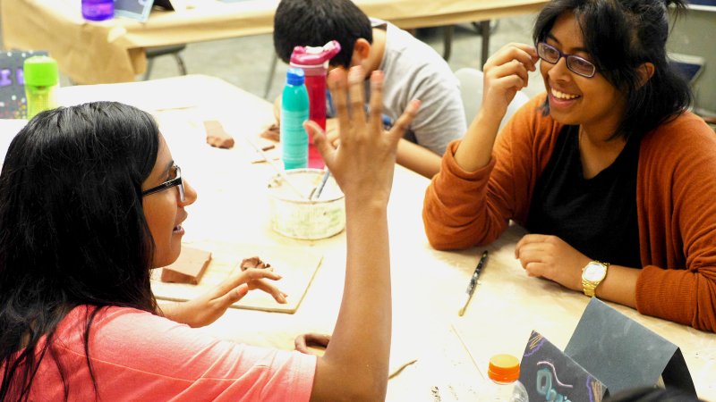 A volunteer smiles as she listens to a child talk while another child sits nearby, immersed in an art activity.