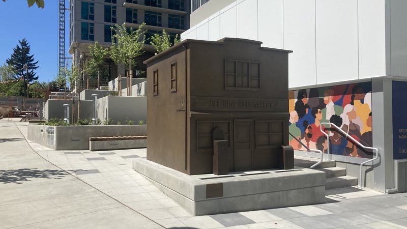 A bronze sculpture of a small building labeled 'Cheung's Food Market' in an urban plaza, with a colorful mural in the background.