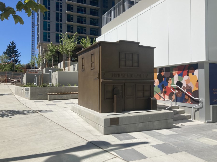 A bronze sculpture of a small building labeled 'Cheung's Food Market' in an urban plaza, with a colorful mural in the background.