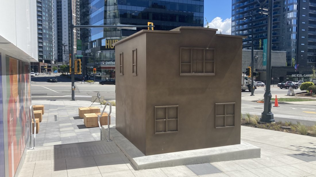 A bronze sculpture of a small building labeled 'Cheung's Food Market' in an urban plaza, with a colorful mural in the background.