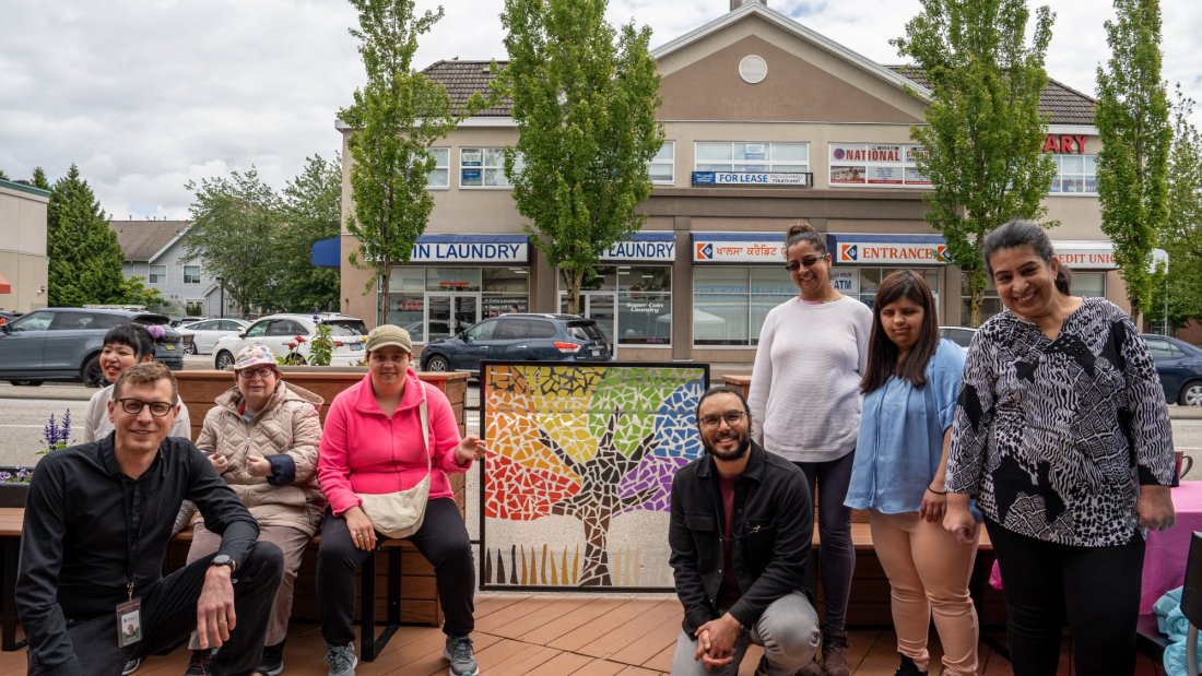 Diverse group of people smiling, gathered around a colorful mosaic artwork on an outdoor urban patio with wooden benches and planters.