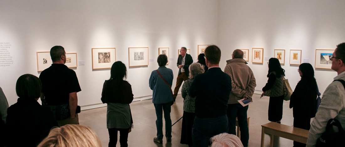 A gathering of people from the back in a white gallery space, looking at framed artworks on the wall.