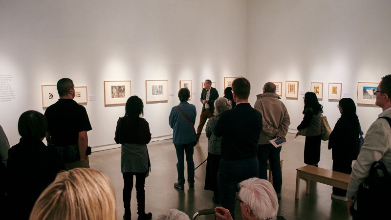 A gathering of people from the back in a white gallery space, looking at framed artworks on the wall.
