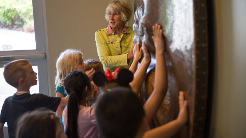 Docent Volunteer Ev Zaklan looks at a group of schoolchildren as they touch an artwork that resembles a mandala and that can be spun around.