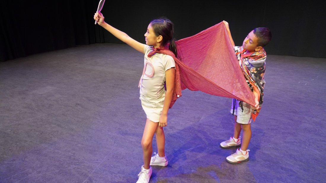 young girl looking into a hand mirror, while young boy holds her red cape spread out behind her