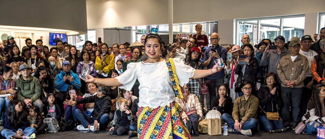 Filipino female dancer in traditional attire performing at an indoor community event with a large, diverse audience watching and taking photos.