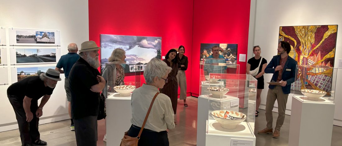 A group of people stand amongst plinths of artworks and paintings in a gallery painted white and red as a curator gives a tour.