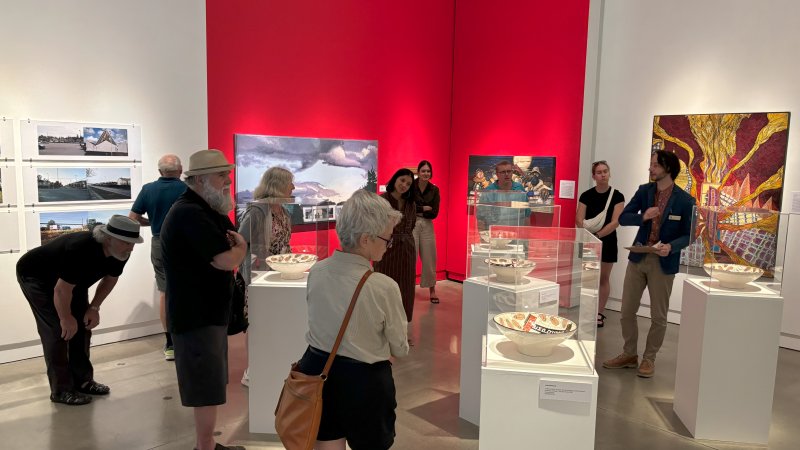 A group of people stand amongst plinths of artworks and paintings in a gallery painted white and red as a curator gives a tour.
