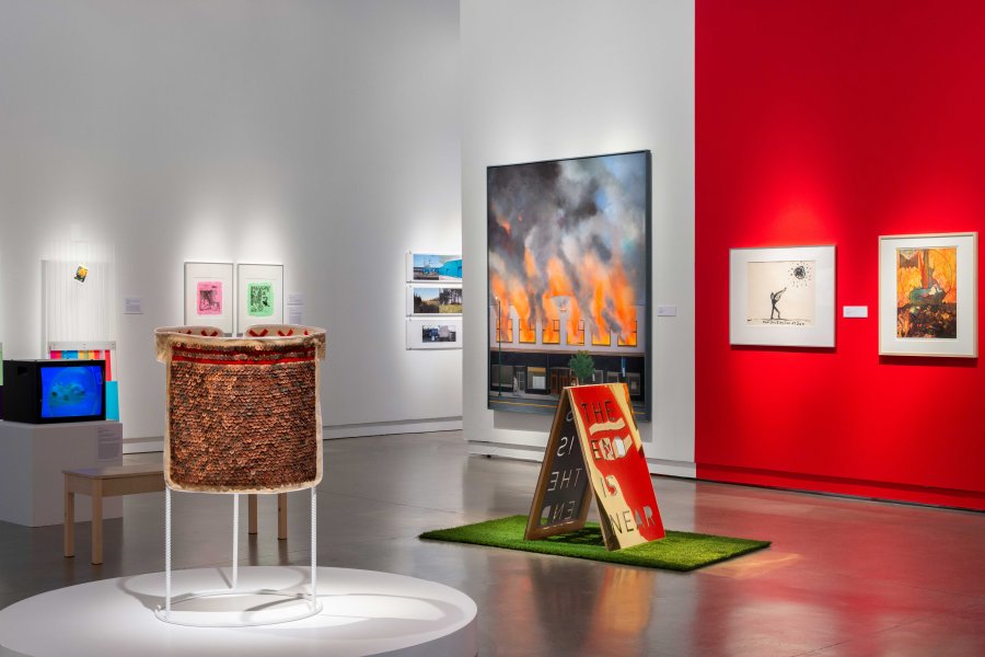 Various artworks in a gallery space with white and red walls: a sandwich board with the phrase "the end is near" on it, a sculpture of pennies on an apron, and paintings, one of which shows a storefront on fire.