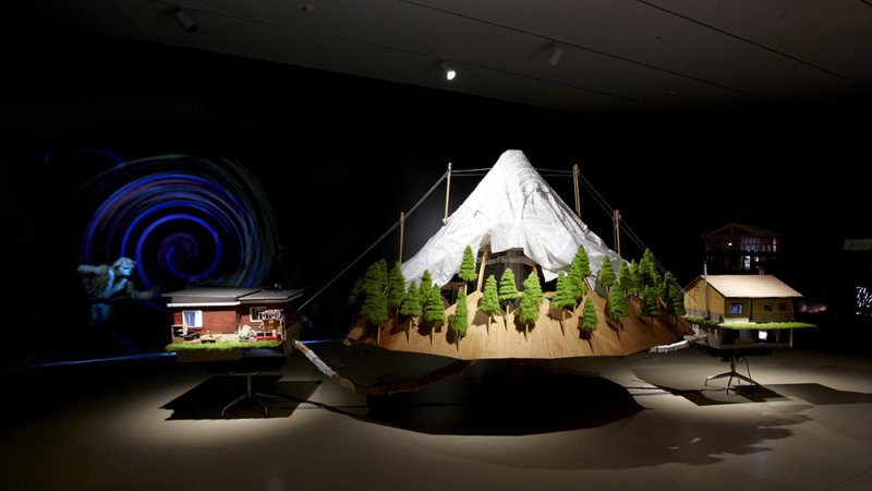 A snowy mountain with coniferous trees at its base appears to float above the ground beside two residential houses that hover from the floor. On the wall is a circular shape made of dark blue and black swirls. There is a figure running out of the swirl. 