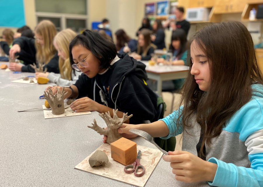 A row of children seated at a table each working on moulding and creating with clay. They are focused on creating tree-like figurines.