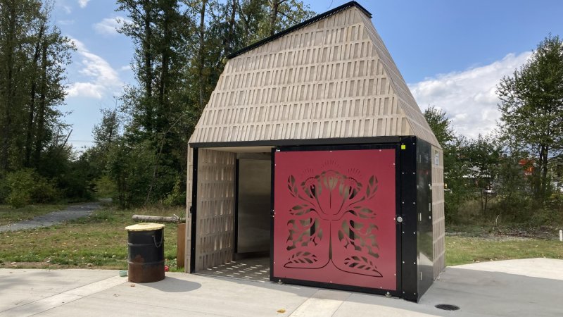 A modern restroom facility in a park with a detailed red tree design on its door, surrounded by trees under a clear sky.
