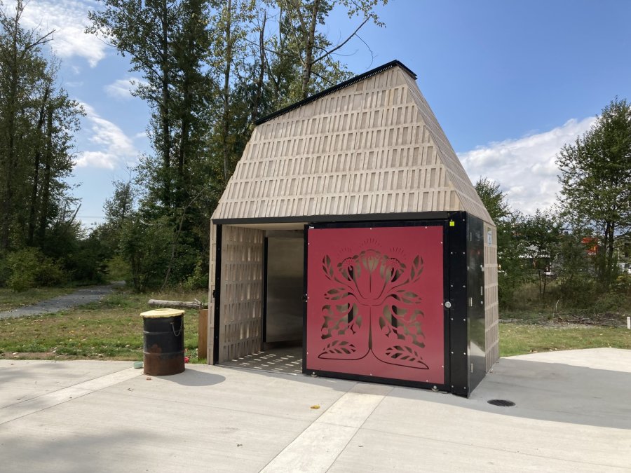A modern restroom facility in a park with a detailed red tree design on its door, surrounded by trees under a clear sky.
