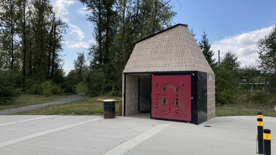 A modern restroom facility in a park with a detailed red tree design on its door, surrounded by trees under a clear sky.