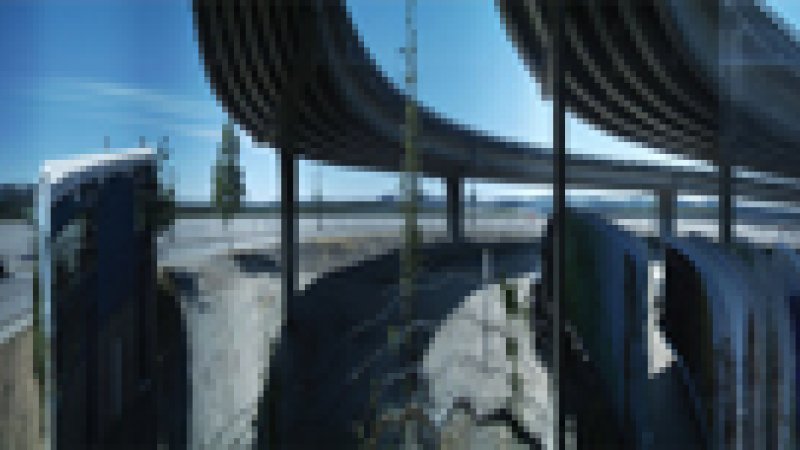 A wide panorama of the urban landscape in metro Vancouver. Taken at one of the SkyTrain station stops, there is a road and two SkyTrain tracks that goes above the road. The sky is blue without any clouds, and the streets are grey. It is a bright, dry-looking stretch of street landscape. 