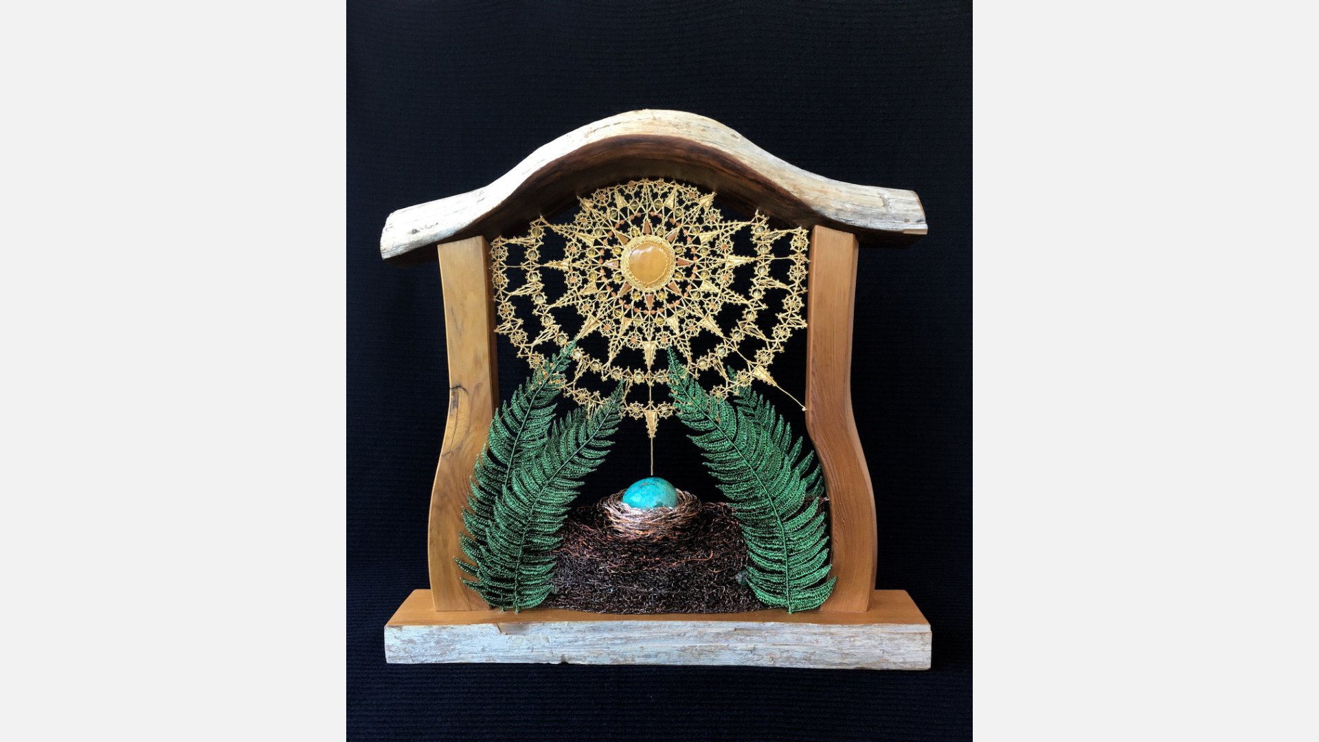 A wooden sculpture housing a nest with a robin's egg at the base, flanked by green ferns. A gold, sun-like shape fills the space above.