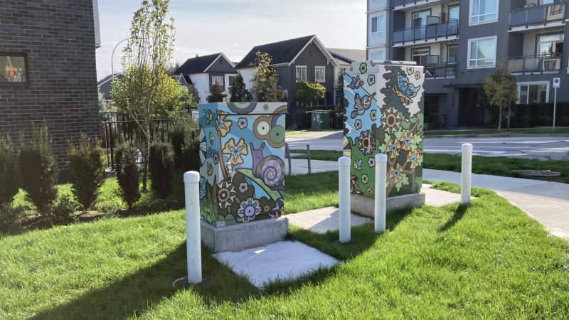 Decorated utility boxes featuring colorful floral and animal-themed artwork, situated in a green space near residential buildings.