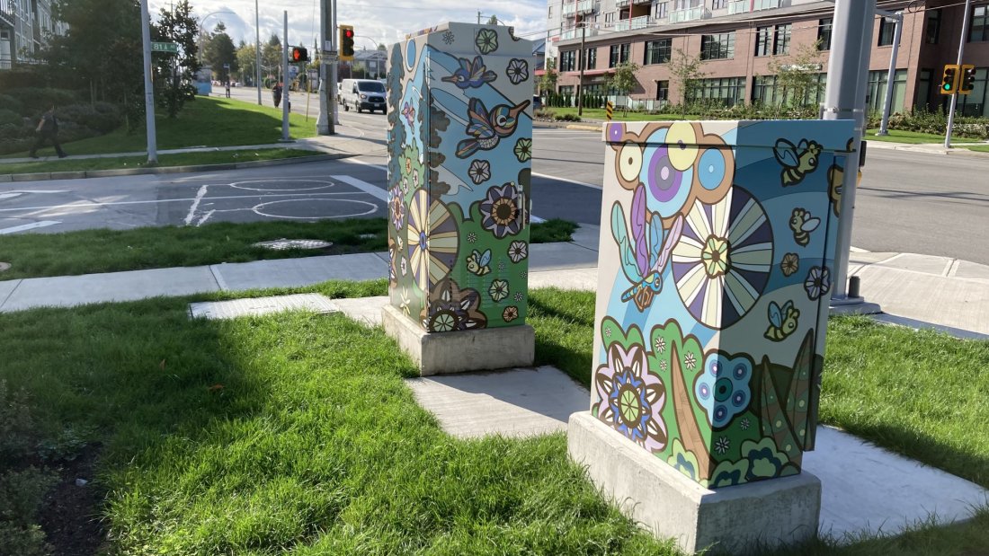 Decorated utility boxes featuring colorful floral and animal-themed artwork, situated in a green space near residential buildings.
