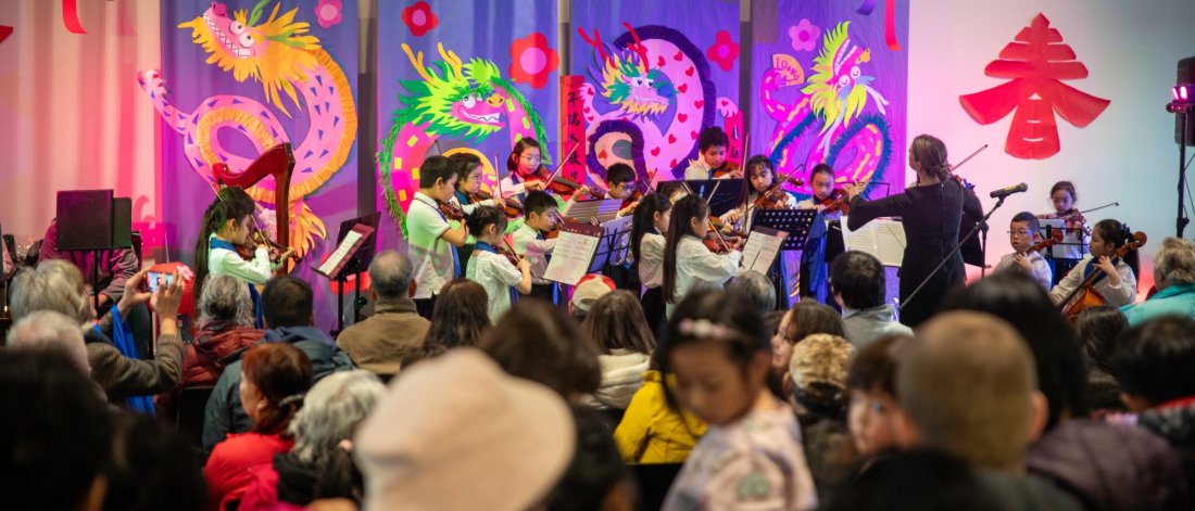 A children's orchestra performing in front of a colorful Lunar New Year-themed backdrop, with an audience seated in the foreground.