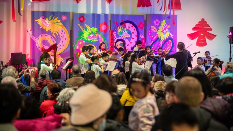 A children's orchestra performing in front of a colorful Lunar New Year-themed backdrop, with an audience seated in the foreground.