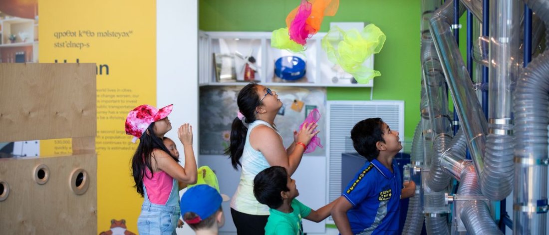 Children engage with interactive exhibits at a museum, playing with colorful floating materials and observing tubing systems