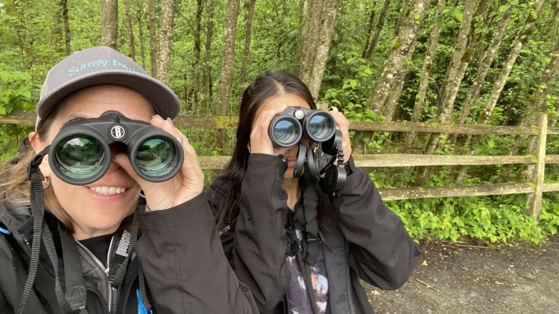Two women using binoculars. Background is of a trail with trees and a wooden fence.