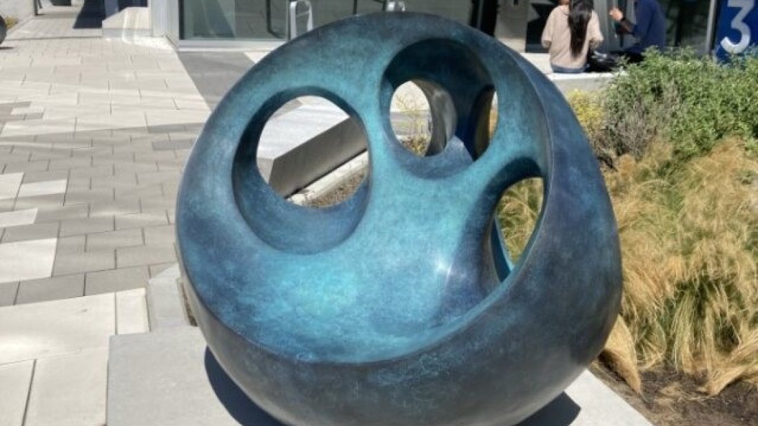  A dark, polished, seed-shaped sculpture with a spiraling groove sits on a wet sidewalk, with bare trees and a street in the background.