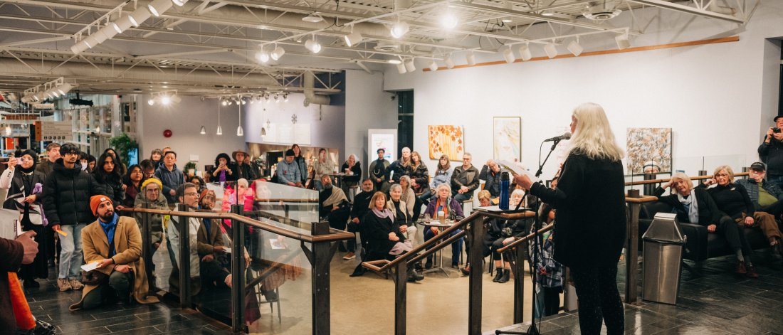 A person stands at the top of a set of stairs, holding papers in her left hand and speaks into a microphone to a crowd of people.