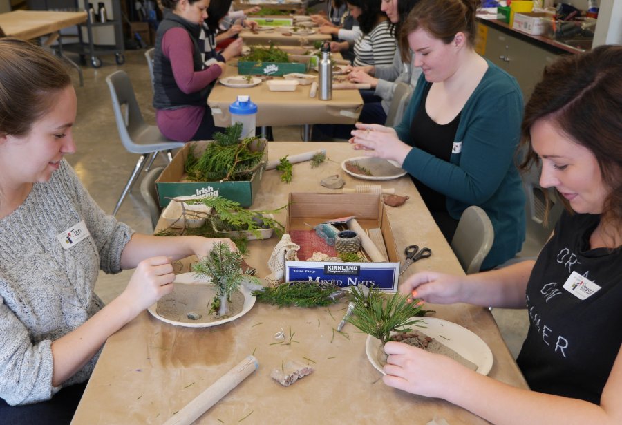 A group of teachers are sitting at tables in a classroom, making creations out of tree branches, rocks, sand, and other natural materials.