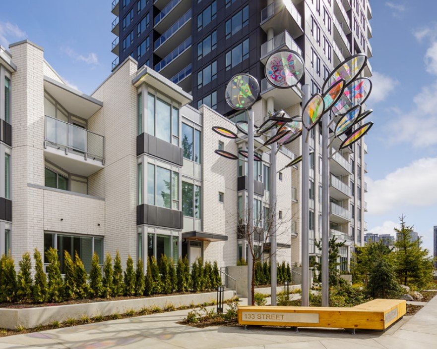A vibrant kinetic sculpture with colorful, translucent elements stands in front of a modern apartment building
