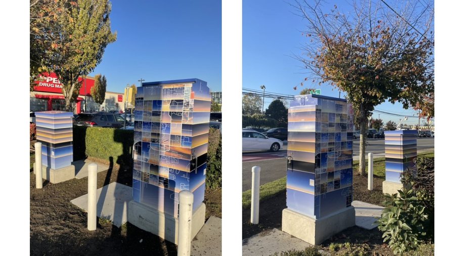 Utility boxes featuring a blue and silver geometric design are positioned on a sidewalk with a tree and a clear sky above.