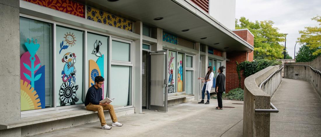 Colourful mural by Sandeep Johal on Surrey Arts Centre classroom windows, with 3 people nearby.