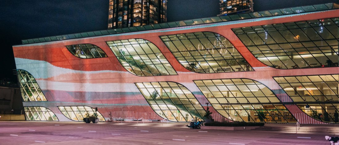 Waves of red, orange, and pale blue are projected on the facade of Surrey City Centre Library whose windows are lit up. A skyscraper stands in the distance behind.
