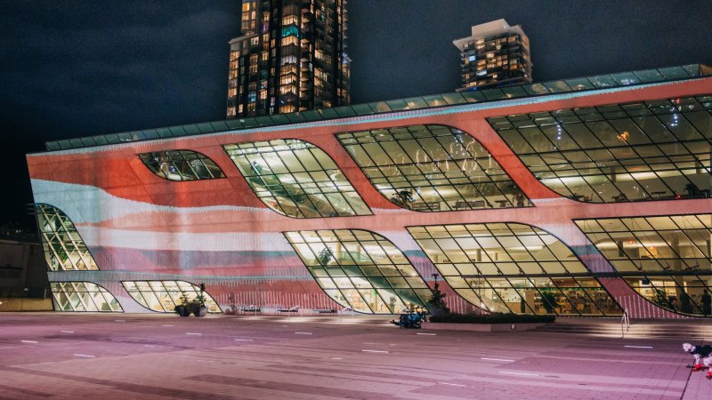 Waves of red, orange, and pale blue are projected on the facade of Surrey City Centre Library whose windows are lit up. A skyscraper stands in the distance behind.