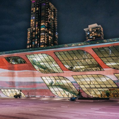 Waves of red, orange, and pale blue are projected on the facade of Surrey City Centre Library whose windows are lit up. A skyscraper stands in the distance behind.