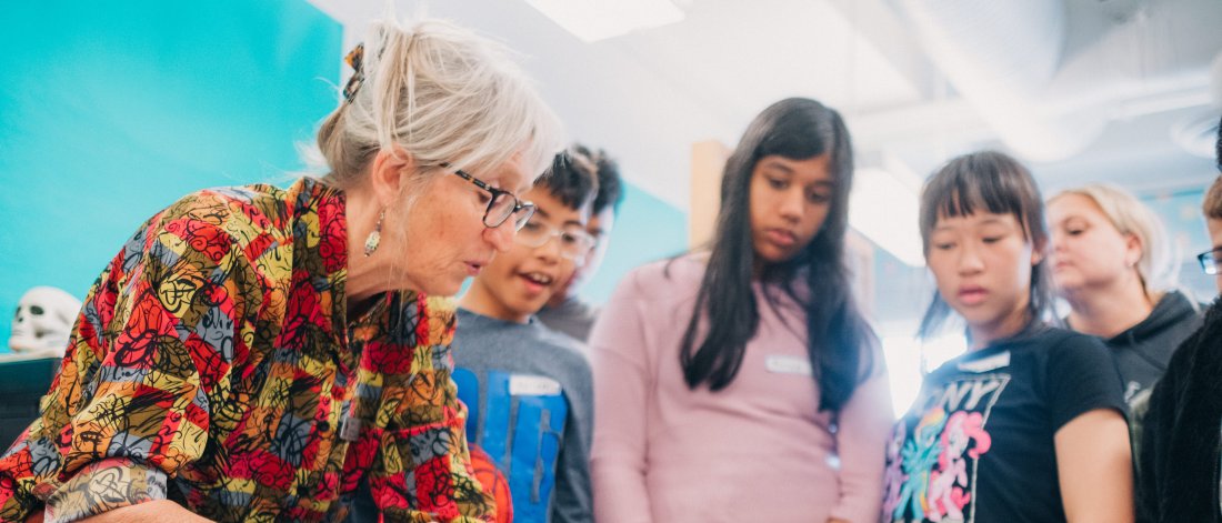 A teacher shows a group of students an artwork print on a table.
