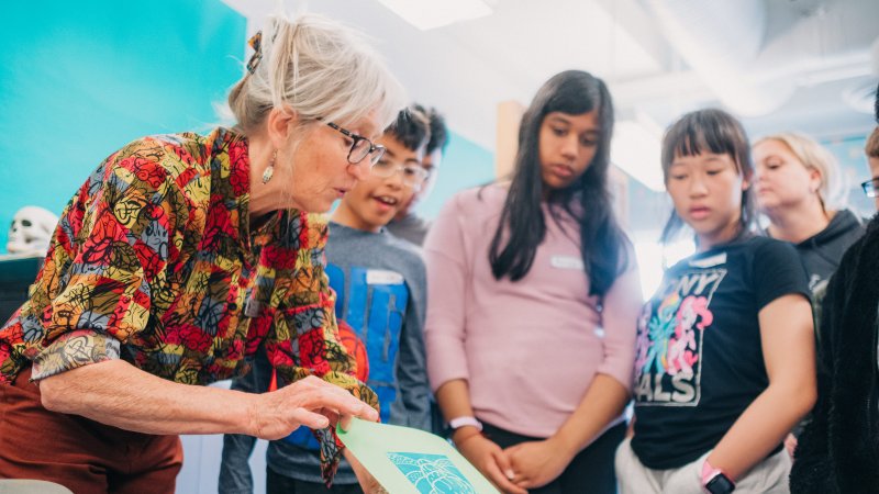 A teacher shows a group of students an artwork print on a table.