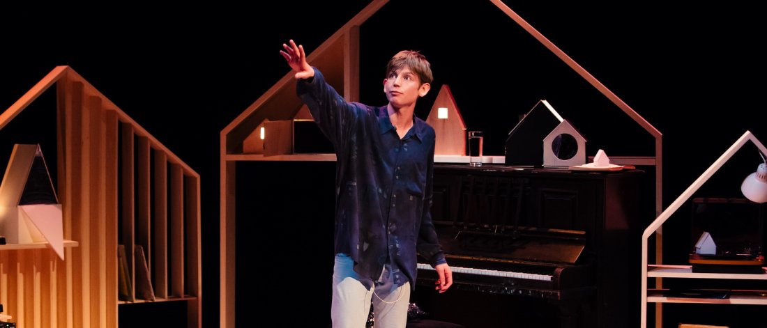 Performer Toby Thompson on stage arm raised in front of a set that includes a piano and wooden pieces that resemble the inside of a house 