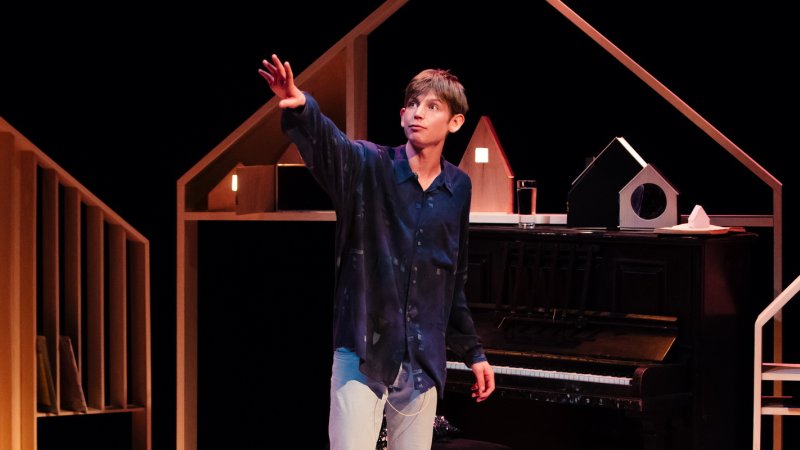 Performer Toby Thompson on stage arm raised in front of a set that includes a piano and wooden pieces that resemble the inside of a house 