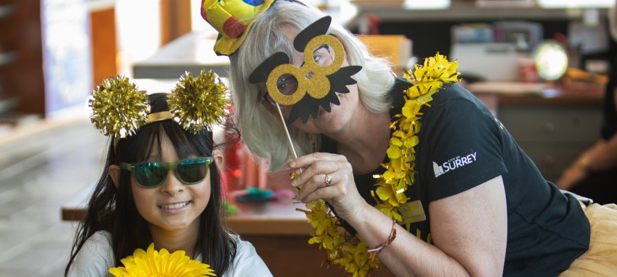 a child wearing sunglasses and a glittery gold pom pom headband and a woman holding up a photo prop of gold glasses and a moustache pose for the camera