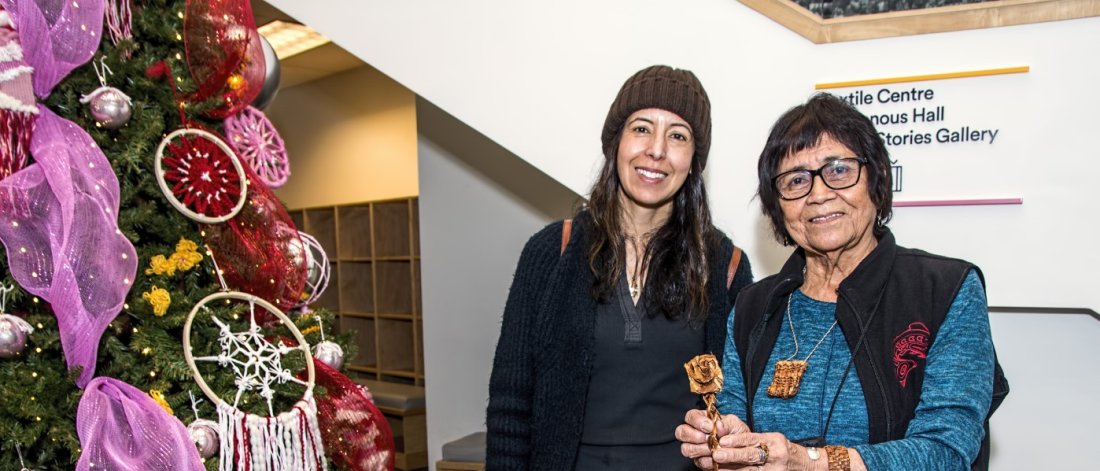 Two women stand smiling behind a display table at a craft market, with a Christmas tree in the background. 